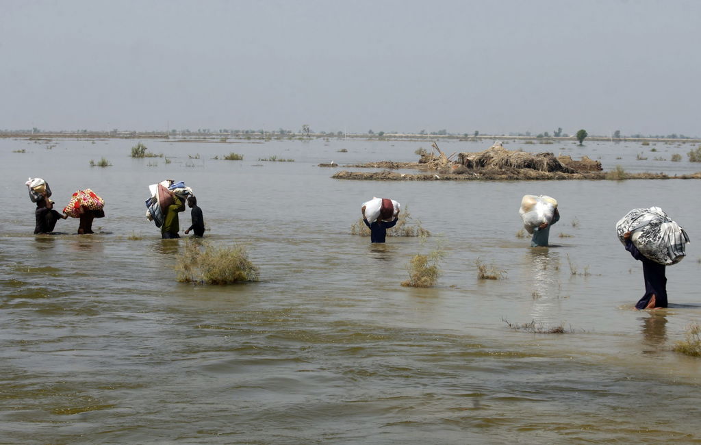 Five people wade through knee-deep water toward the shoreline carrying packages above their heads
