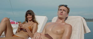 A bikini clad woman with long blonde hair sits on a lounge chair smiling next to a shirtless blonde-haired man who looks perturbed with the ocean and the blue sky visible behind them.